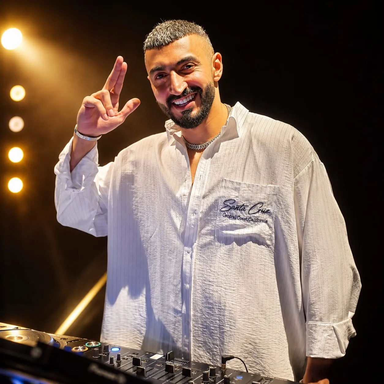 DJ in a loose white shirt giving a peace sign with bulbs glowing behind him