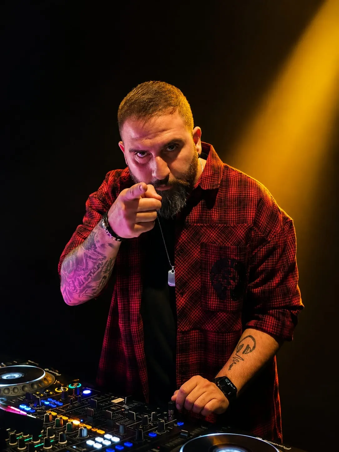 DJ in a red plaid shirt pointing at the camera from behind the mixer